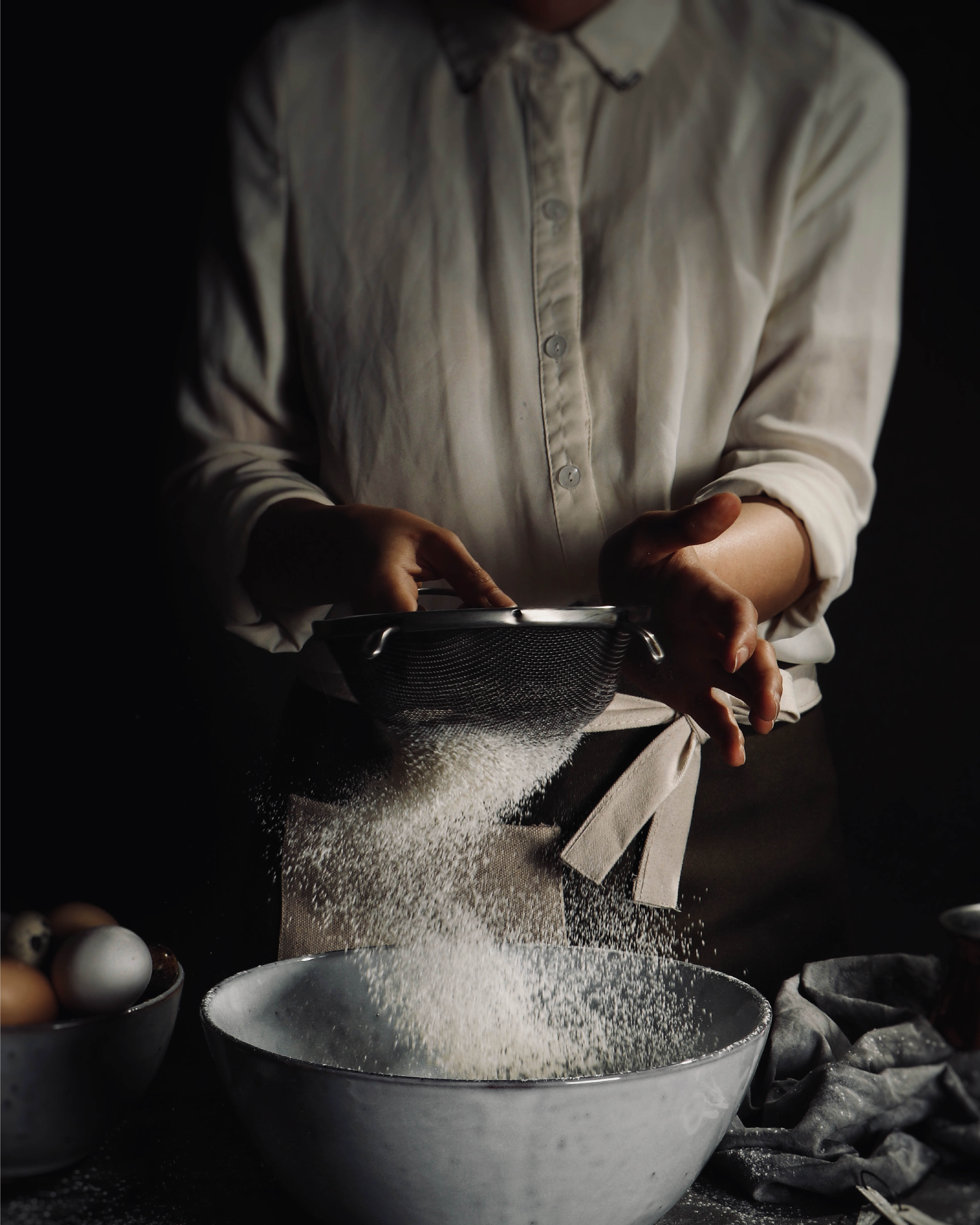 Ambachtelijke bakker zeeft bloem in een kom bij zacht licht, omringd door verse ingrediënten en linnen doeken.