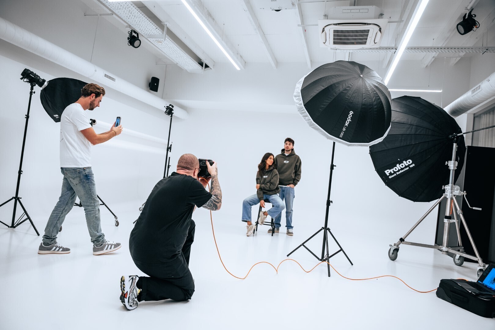 Professionele fotoshoot in een witte studio met fotografen en een koppel dat poseert tussen grote studiolampen.