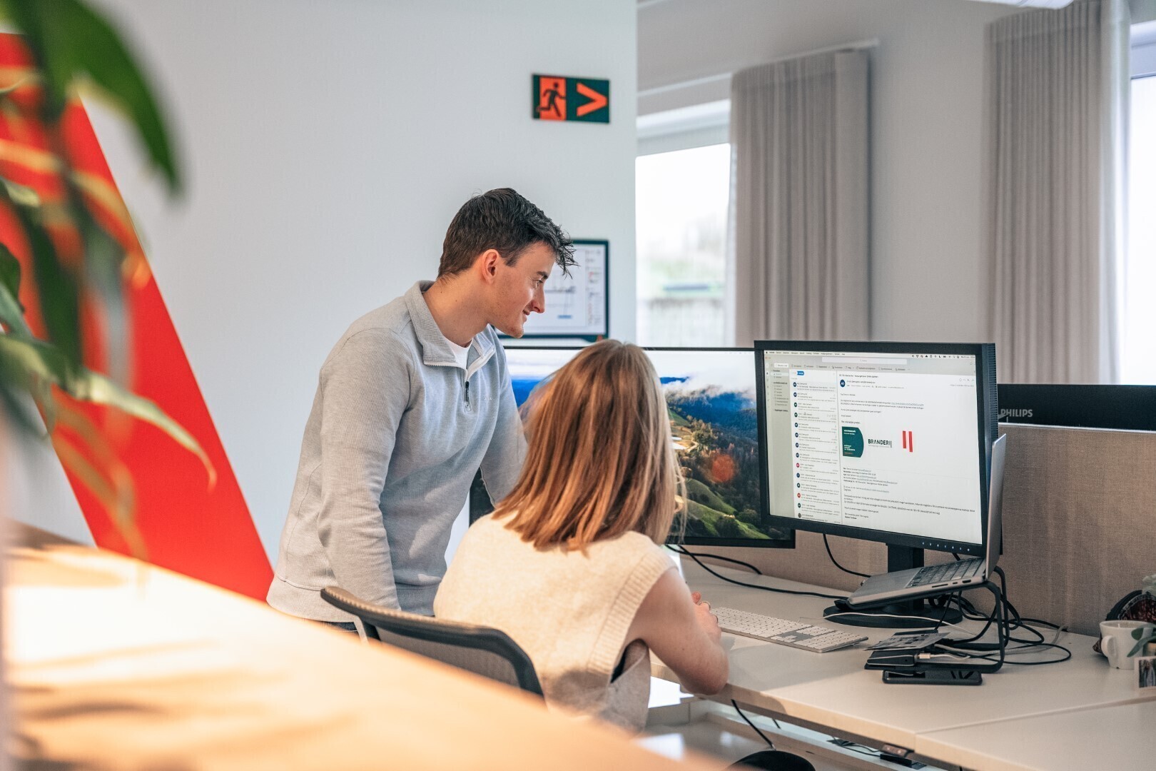 Man en vrouw werken samen aan een dubbele computerset-up in een kantoorruimte, waarbij ze samen naar het scherm kijken en iets bespreken