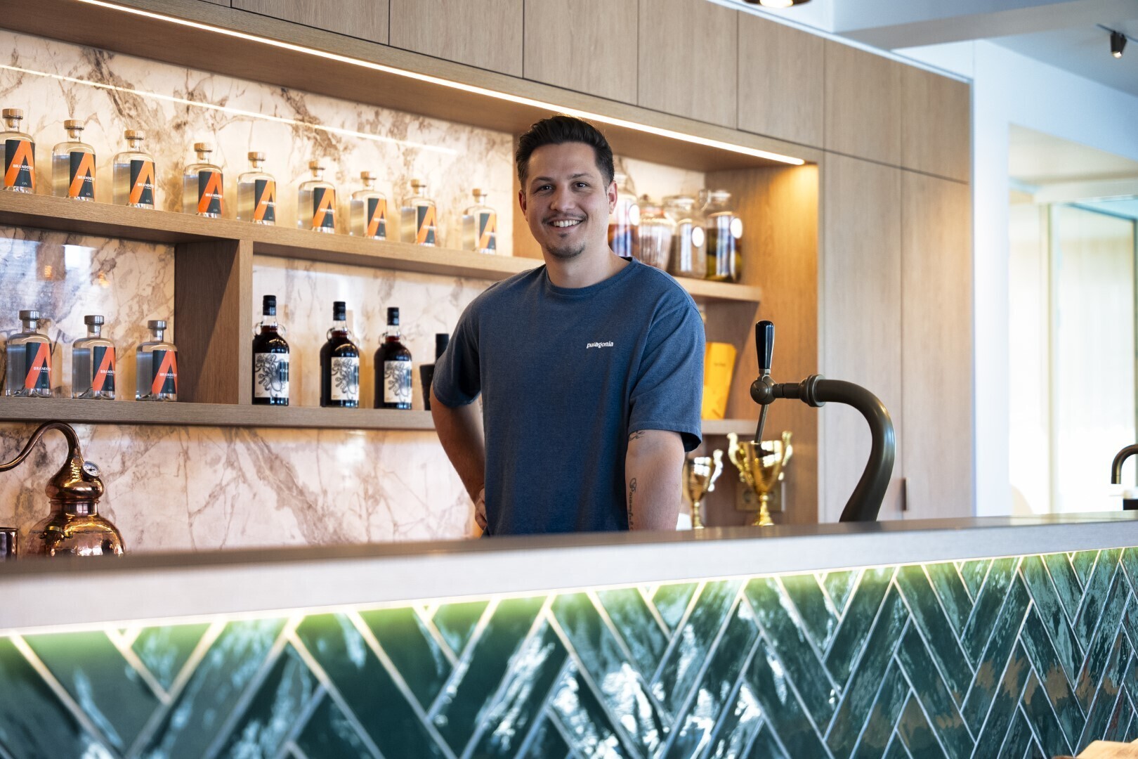 Man in blauw T-shirt staat glimlachend achter een moderne bar met groene tegels en flessen op de achtergrond.