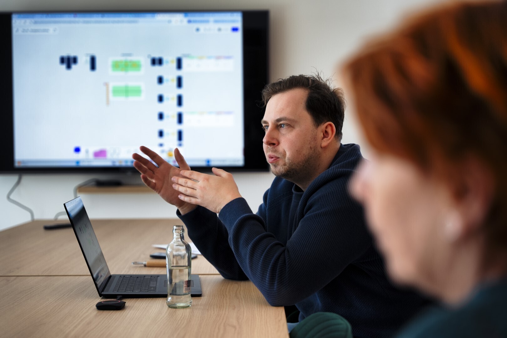 Man spreekt tijdens een strategische sessie aan een vergadertafel, met laptop en flessenwater voor zich, terwijl een planning of flowchart zichtbaar is op het grote scherm achter hem.