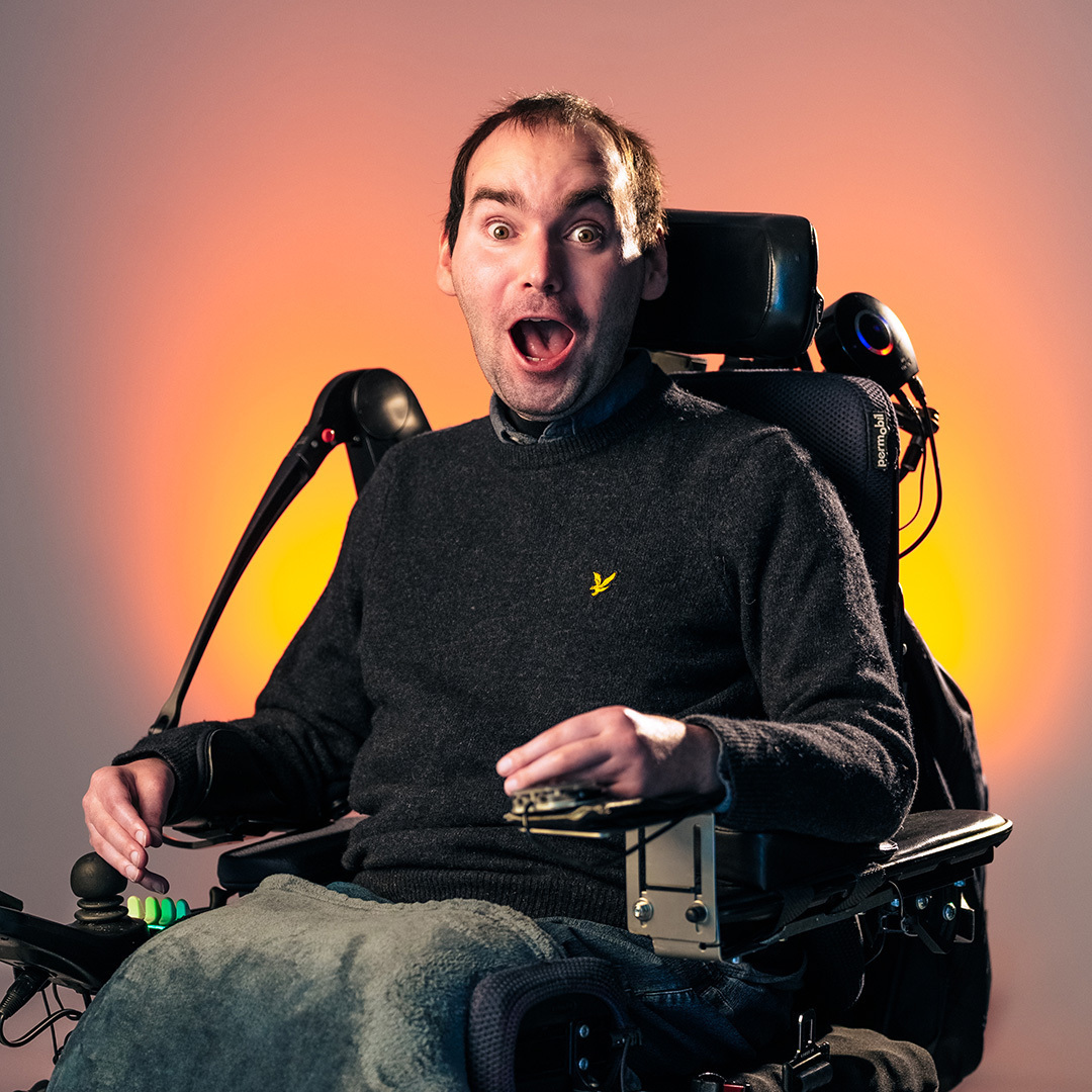 Lachende man in een elektrische rolstoel, gekleed in een donkere trui, poseert voor de camera tegen een neutrale achtergrond