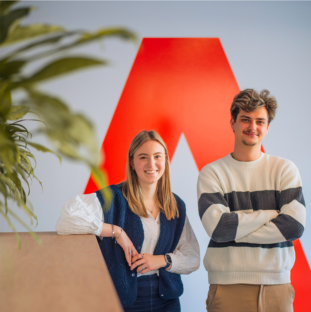 Twee jonge stagairs poseren glimlachend voor een grote rode A-vorm op de muur in een modern kantoor, met groene plant op de voorgrond.